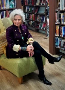 Faith Hunter author full body shot wearing a cool looking purple felt jacket with a military cut over a black shirt, long silver necklace with large turquoise pendant, leather wrist bracelets, black leggings and black shoes, sitting in chair surrounded by book shelves.