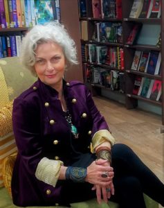 Faith Hunter author close up body shot wearing a cool looking purple felt jacket with a military cut over a black shirt, long turquoise necklace, leather wrist bracelets, sitting in chair surrounded by book shelves.