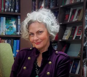 Faith Hunter author closeup upper body head shot wearing a cool looking purple felt jacket with a military cut over a black shirt, long turquoise necklace, sitting in chair surrounded by book shelves.